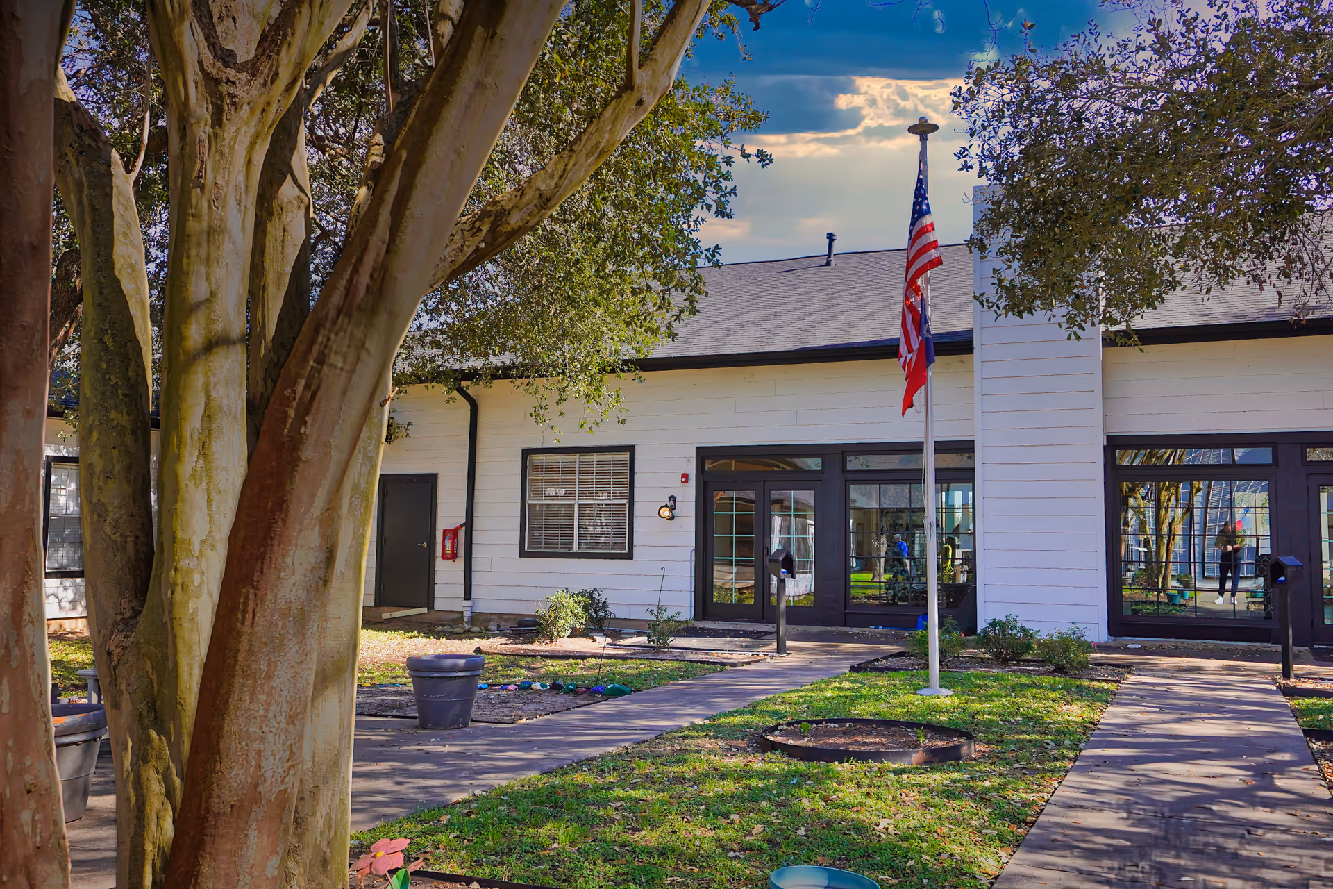 Exterior view of a single-story building with white siding and black trim, featuring a flagpole with an American flag in front. There are trees and a grassy area with a paved walkway leading to the entrance. The building has large windows and a door, with some landscaping around the walkway.