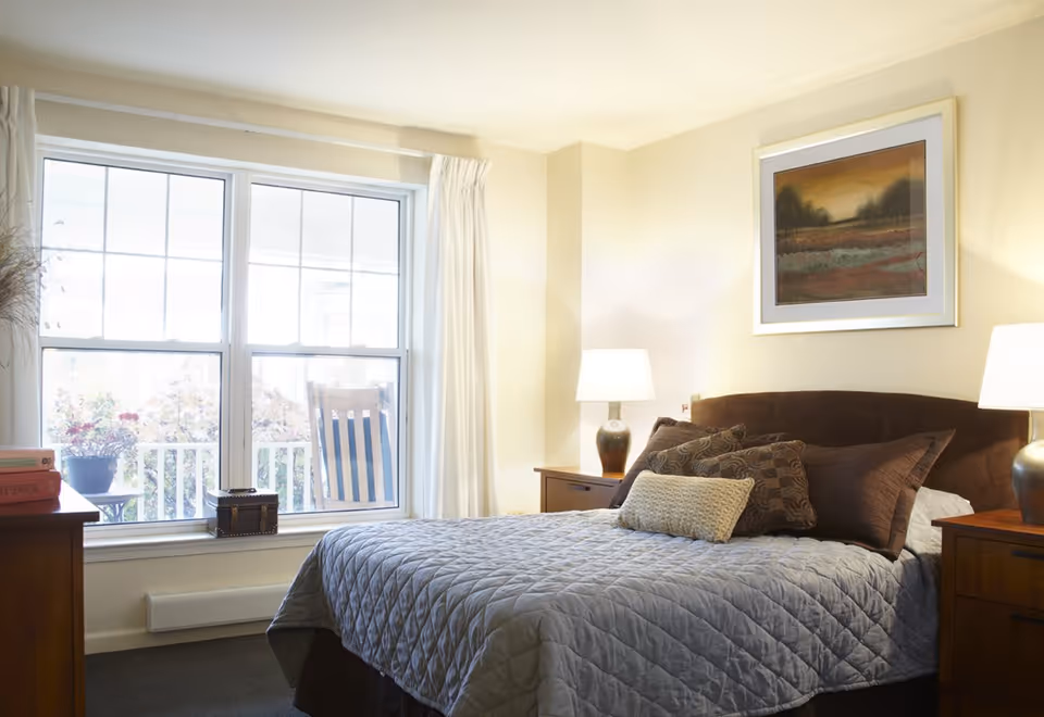 Sunlit bedroom with a neatly made bed, pillows, two bedside tables with lamps, and a large window overlooking a porch.