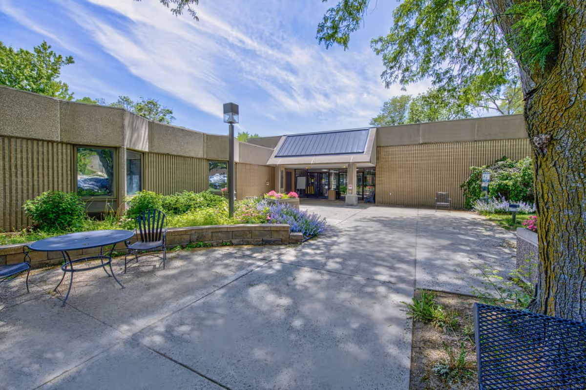 Outdoor view of the entrance to a single-story building with a flat roof and large windows. There are benches and a table with chairs on a concrete patio surrounded by greenery and flowers. Trees provide shade and the sky is partly cloudy.