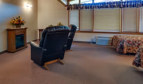 Interior view of a senior living facility room with two black recliner chairs facing a small electric fireplace. The room has carpeted flooring, a large window with blinds and a valance, and a bed with a floral bedspread partially visible on the right side.