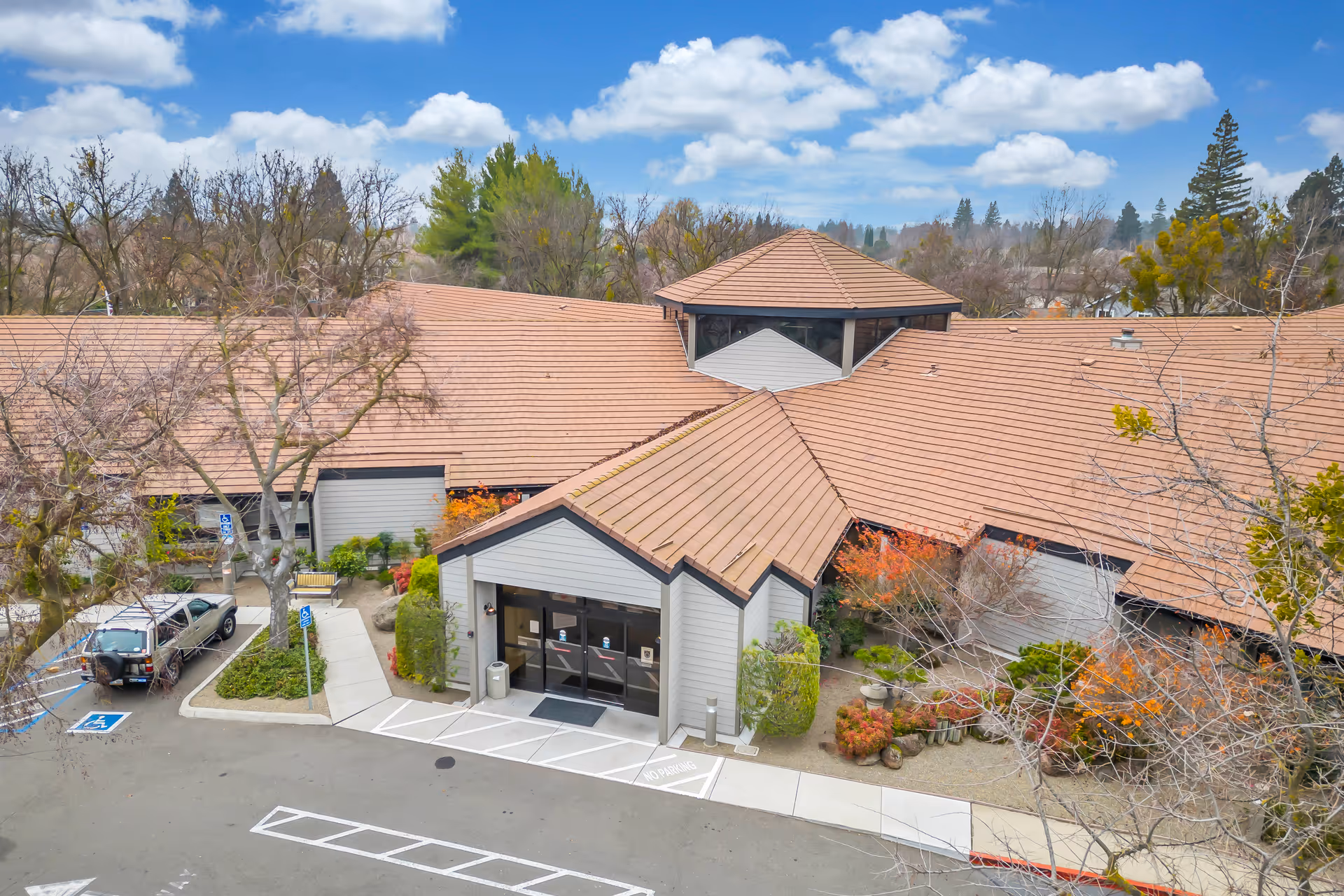 Front exterior aerial view of a single-story care facility with a tan tiled roof, main entrance canopy, landscaped beds, and a few parked cars.