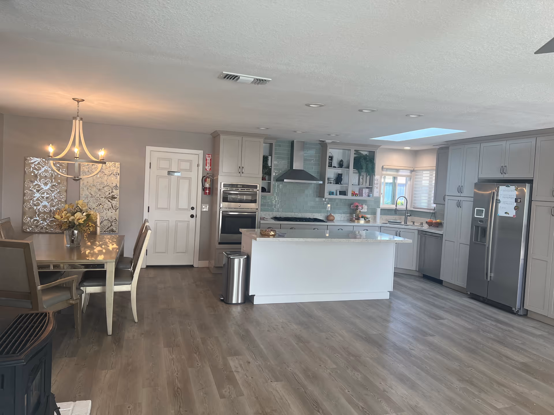 A modern kitchen and dining area with light gray cabinets, a large island with a white countertop, stainless steel appliances including a refrigerator, oven, and dishwasher. The dining area has a wooden table with chairs and a chandelier overhead. The floor is wood, and there is a skylight above the kitchen area.