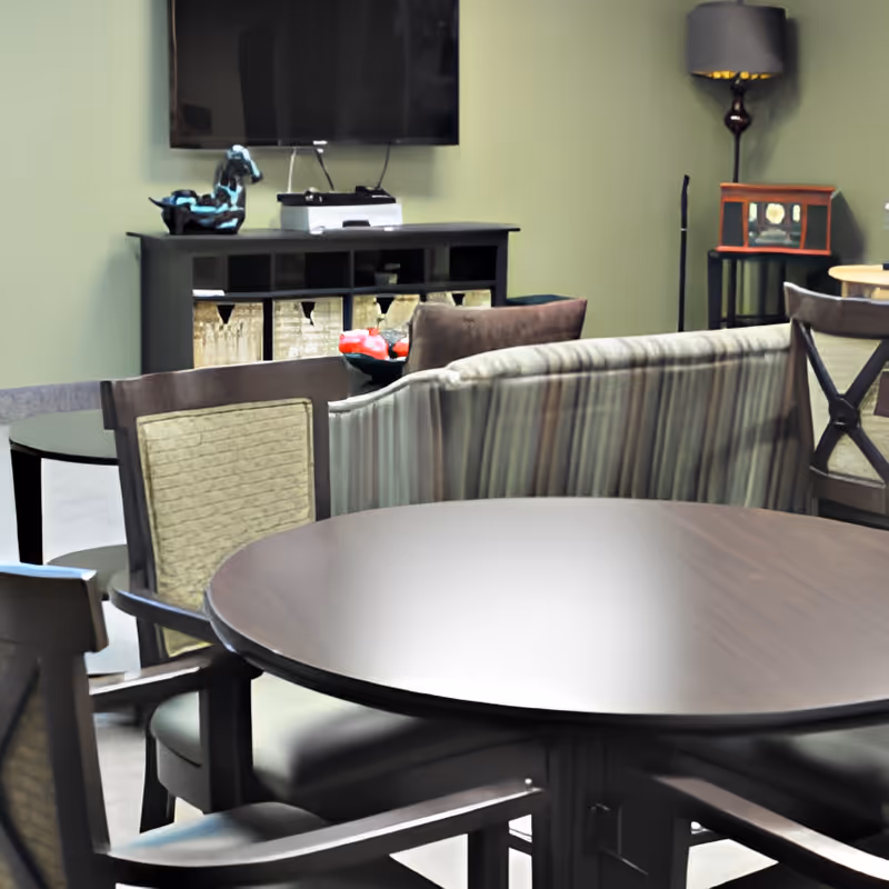 Round dining table and chairs in a communal lounge area with a striped sofa, wall-mounted TV, and storage cabinet.