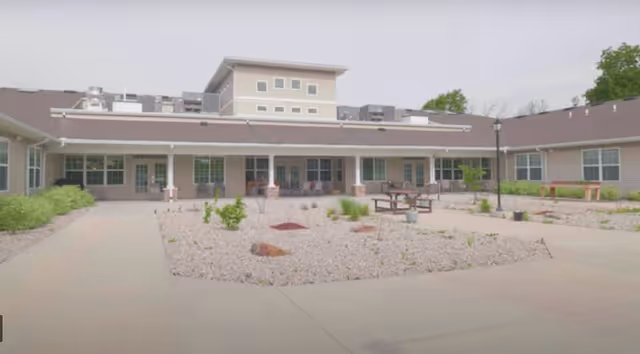 Outdoor courtyard of a senior living facility with gravel landscaping, benches, and a single-story building with a covered walkway and rows of windows.