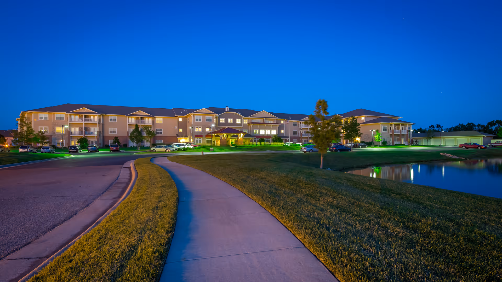 Nighttime exterior view of a three-story senior living building with a curved walkway, lawn, parked cars, and a small reflecting pond.