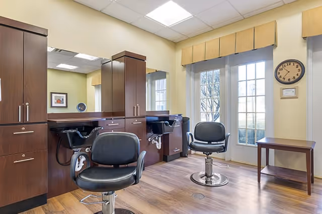 Interior view of a salon area with two black salon chairs in front of wooden cabinets and mirrors. There is a clock on the wall and large windows with glass doors letting in natural light.