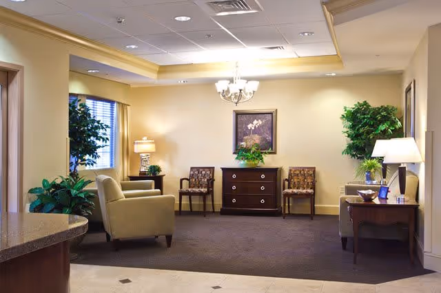 A well-lit interior common area with beige walls and carpeted floor. The room features two armchairs, two patterned chairs, a wooden chest of drawers with a plant and framed artwork above it, and two side tables with lamps. There are also two large potted plants near the windows and walls, and a chandelier hanging from the ceiling.