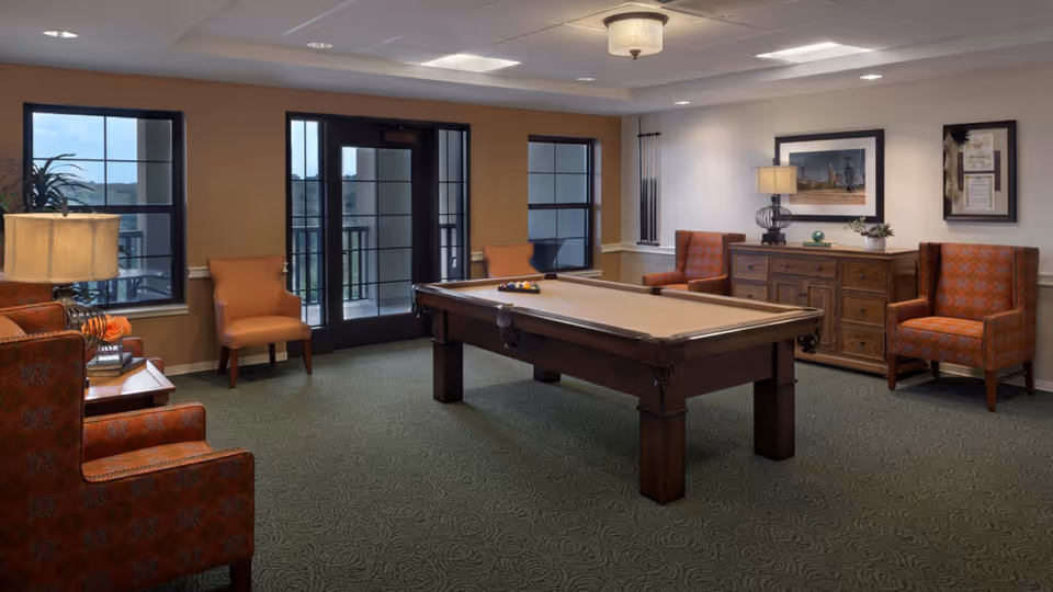 A well-lit recreational room with a pool table in the center, surrounded by several upholstered chairs in orange and patterned fabric. There is a wooden sideboard against the wall with a lamp and framed pictures above it. Large windows and a glass door provide natural light and a view of the outdoors.