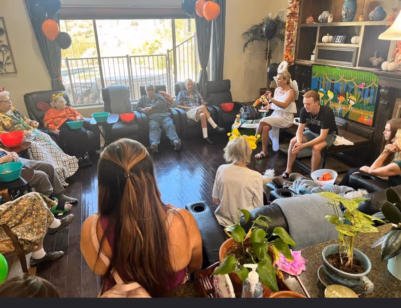 A group of elderly people and caregivers sitting in a circle in a cozy living room with dark wooden floors, large windows with curtains, and a fireplace decorated with autumn-themed items and balloons. Some people are holding bowls, and there are plants and a hand sanitizer bottle on a nearby table.