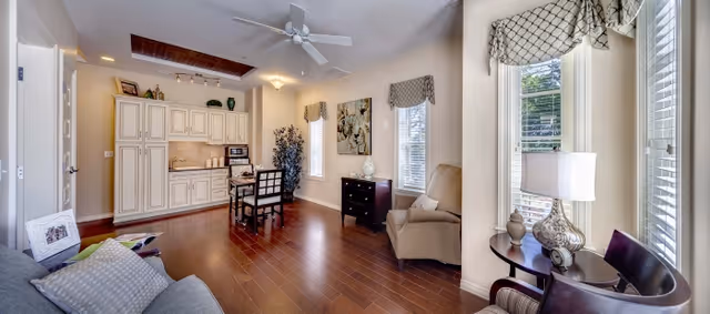 A bright and spacious living area with hardwood floors, featuring a small dining table with chairs near a kitchenette with white cabinets. The room has large windows with patterned valances, a beige armchair, a dark wood side table with a lamp, and a gray sofa with cushions. Ceiling fan and decorative plants and artwork complete the cozy interior.