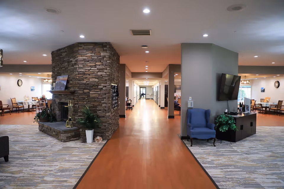 Spacious assisted-living common area with a central stone fireplace, seating, TV, and a long hallway leading to resident rooms.
