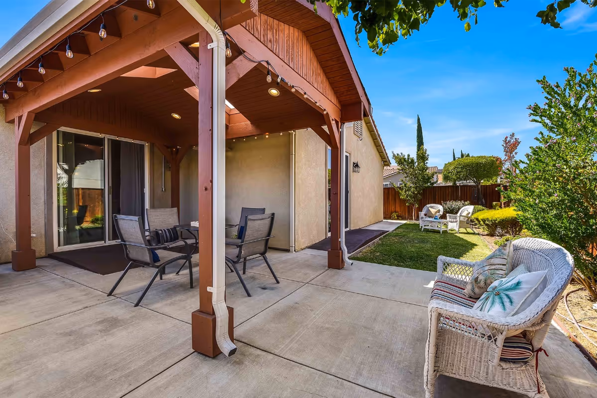 Outdoor patio area with a wooden pergola covering a seating area that includes a glass table and four chairs. Adjacent to the patio is a small grassy yard with additional white wicker seating and various bushes and trees under a clear blue sky.