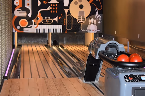 A small indoor bowling alley lane with a colorful music-themed mural at the end. The lane has wooden flooring and a bowling ball return machine holding three red bowling balls.
