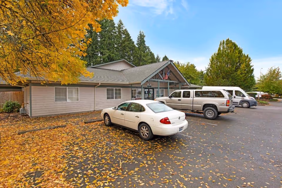 Exterior view of a single-story building with a parking lot in front. Several vehicles are parked, and the ground is covered with fallen autumn leaves. Trees with yellow and green foliage surround the area under a clear blue sky.