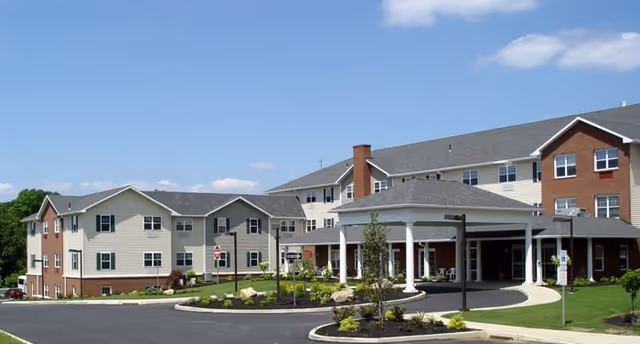 Exterior view of Traditions of Hanover senior living facility showing a large multi-story building with beige and brick siding, a covered entrance with white columns, landscaped grounds, and a clear blue sky.