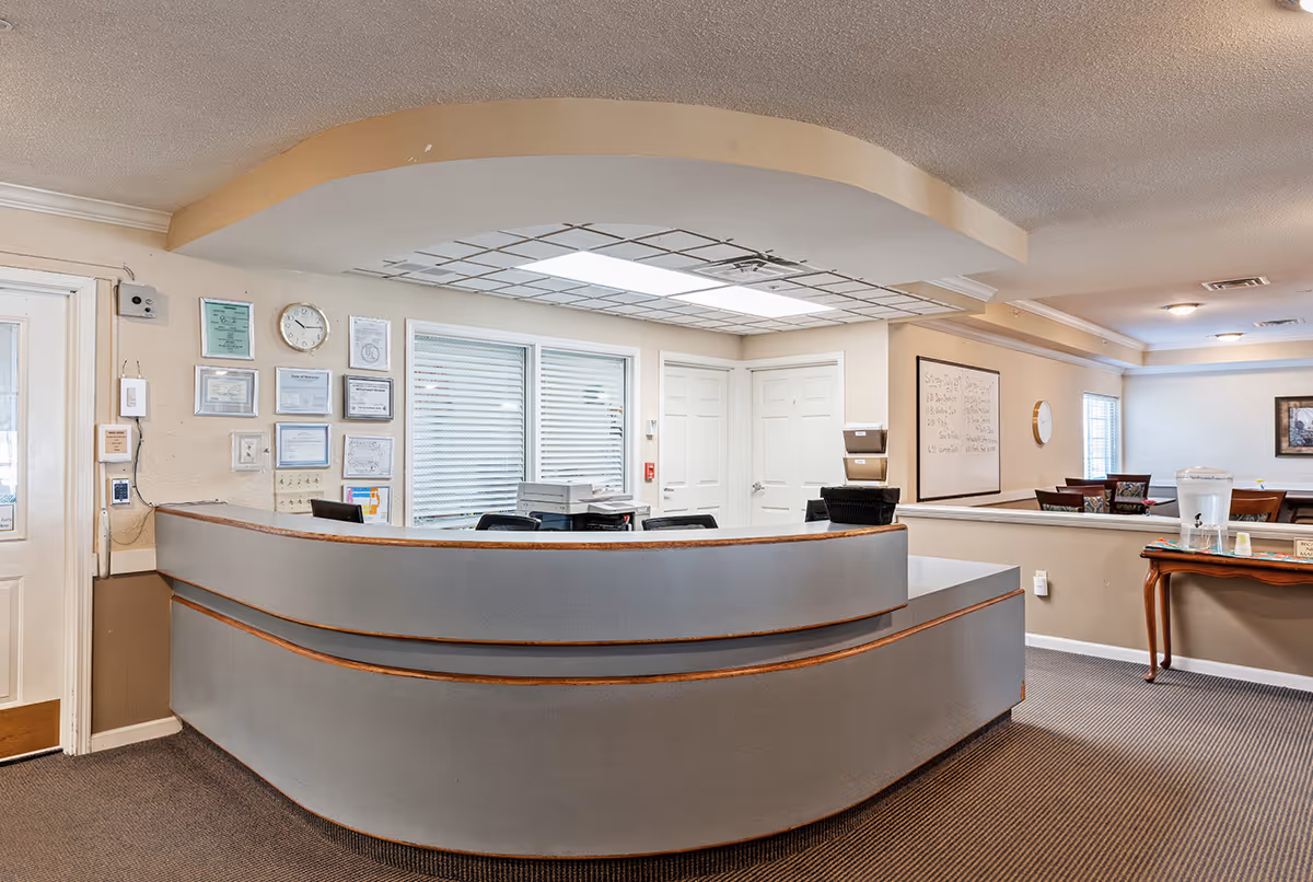 Curved reception desk and lobby area of a senior living facility with certificates on the wall and a seating/dining area beyond.