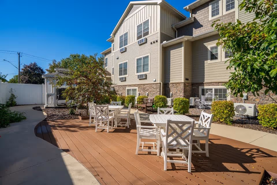 Outdoor patio area at The Harbors Senior Living of Fridley featuring white tables and chairs on a wooden deck, surrounded by shrubs and trees, with a multi-story beige building in the background under a clear blue sky.