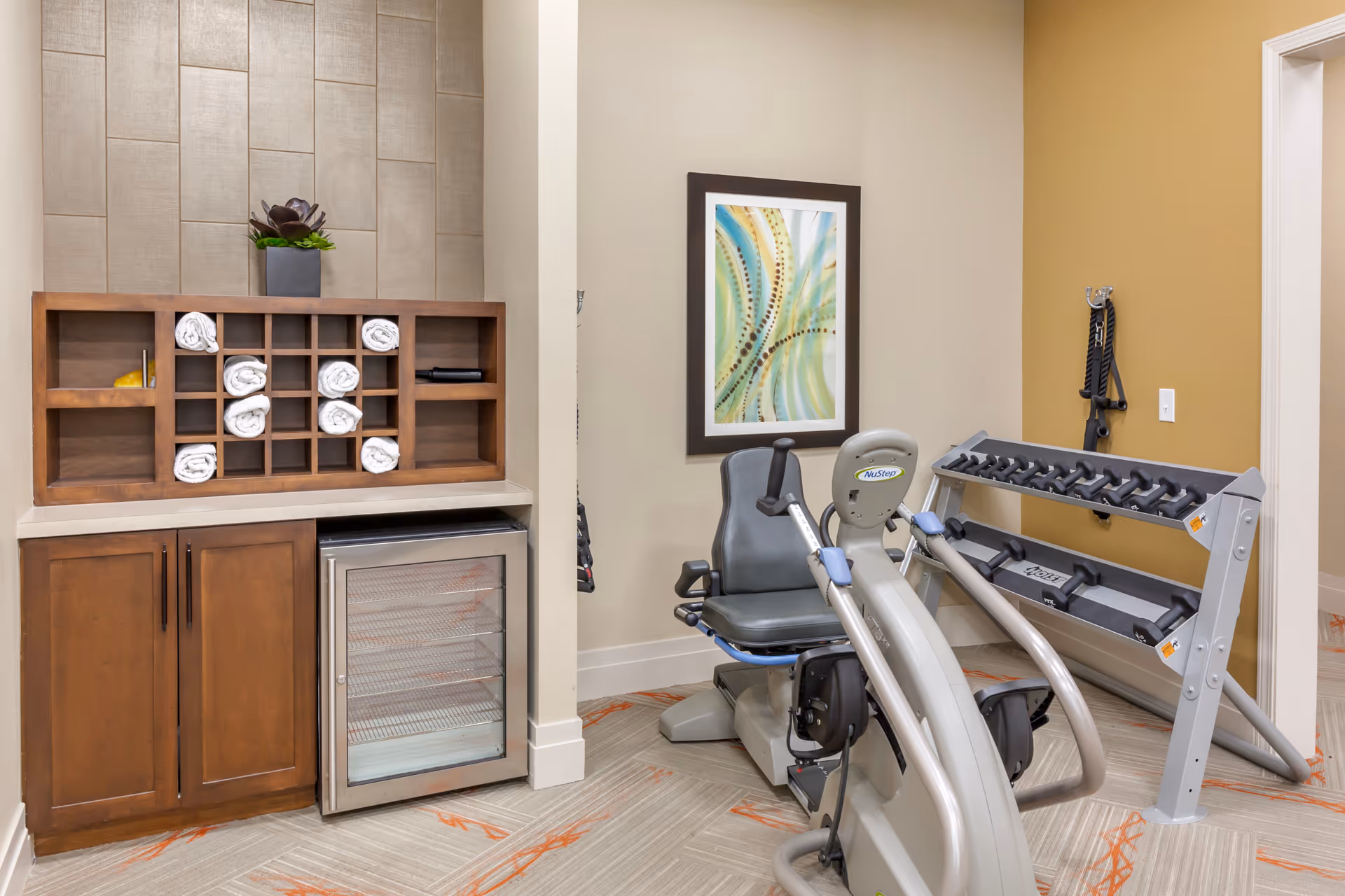 A small fitness room with a recumbent exercise bike, a rack of dumbbells, and resistance bands hanging on the wall. There is a wooden cabinet with rolled white towels and a small refrigerator below. A framed abstract artwork is hung on the wall.