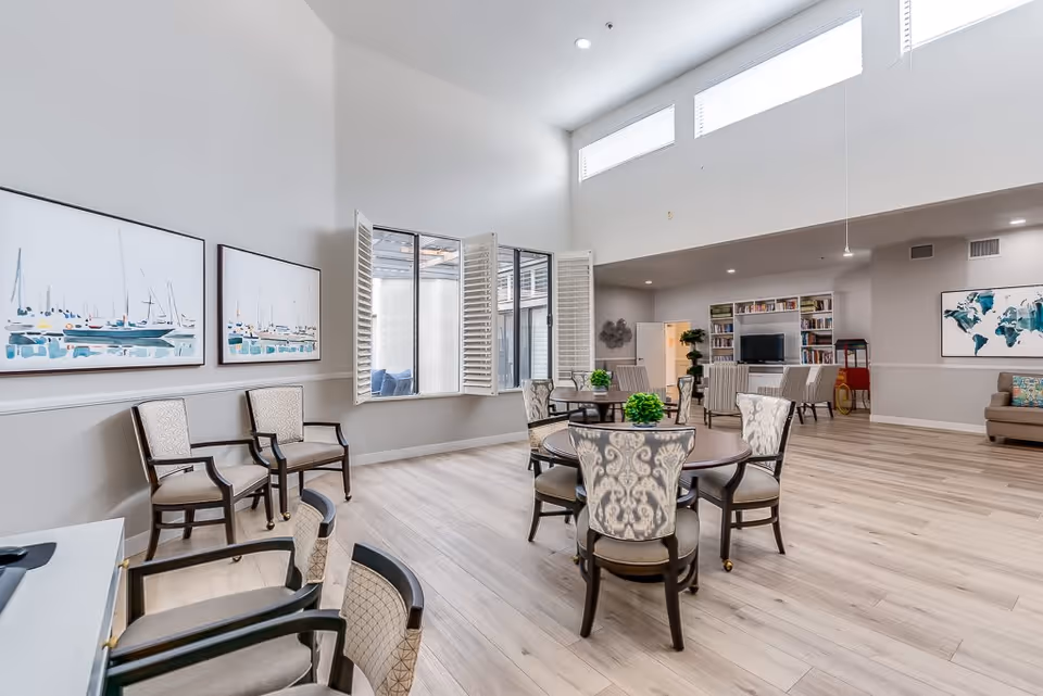 A spacious senior living common area with high ceilings and large windows. The room features several round tables with patterned chairs, additional seating along the walls, light wood flooring, and wall art including paintings of boats and a world map. There is a TV and bookshelf in the background, and the space is well-lit with natural and recessed lighting.