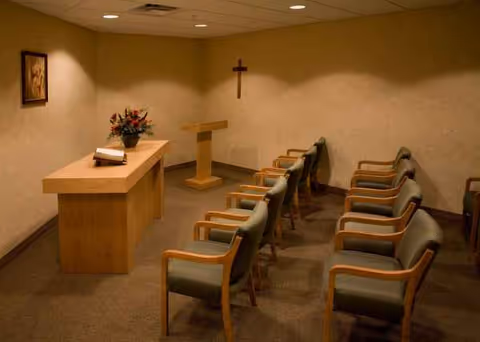 Chapel-like interior with a lectern, cross on the wall, flowers, and rows of chairs.