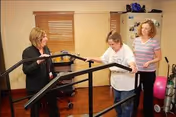 Three women in a room with wooden floors and beige walls. One woman is using a parallel bar walking aid while the other two women assist and support her. There is exercise equipment and a pink exercise ball in the background.