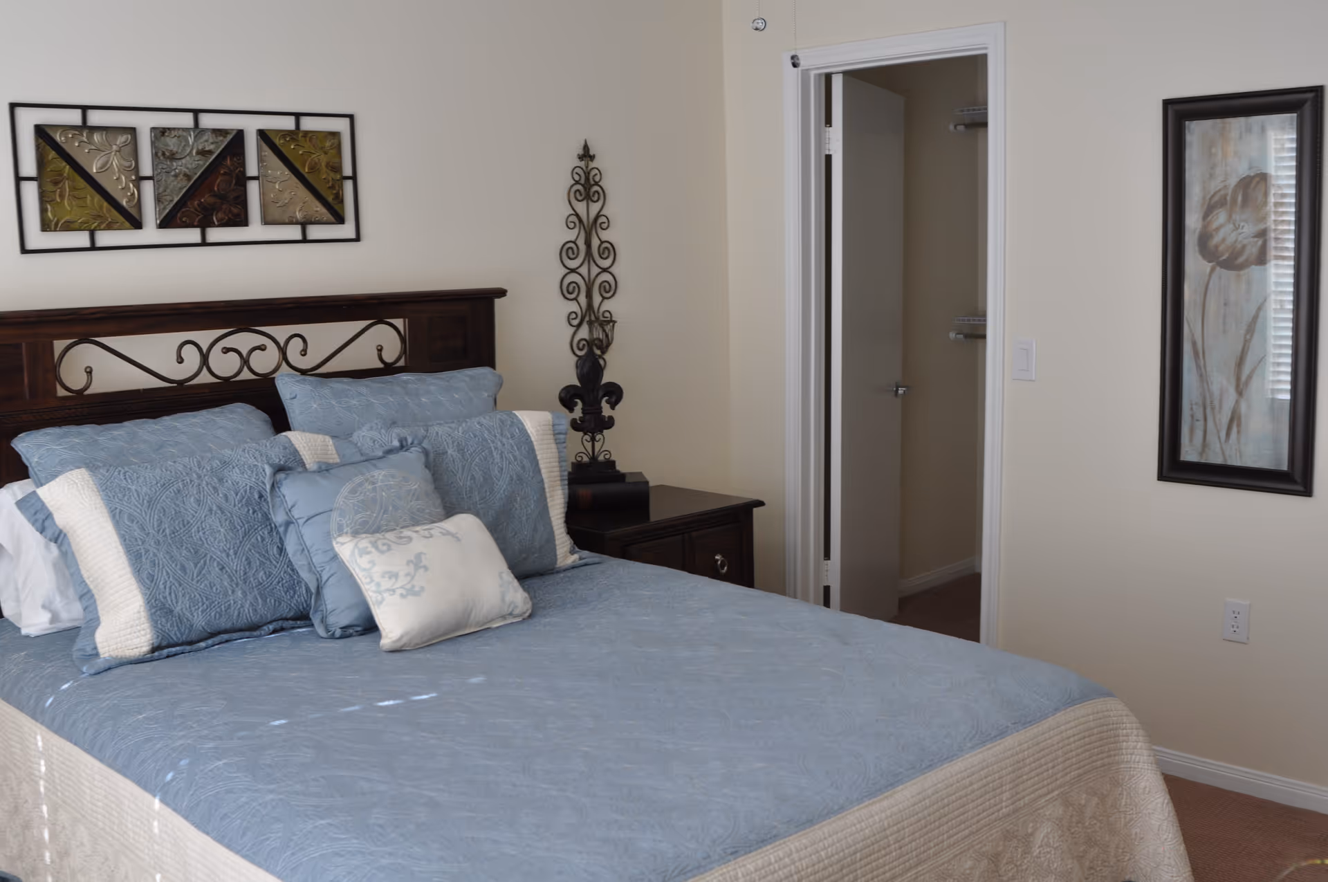 A neatly made bed with blue and beige bedding and multiple pillows in a bedroom. The bed has a dark wooden headboard with decorative metalwork. There is a nightstand with a decorative black metal ornament and a small black box. On the wall above the bed is a framed abstract artwork with various colors. Another framed floral artwork hangs on the adjacent wall near an open doorway leading to another room.
