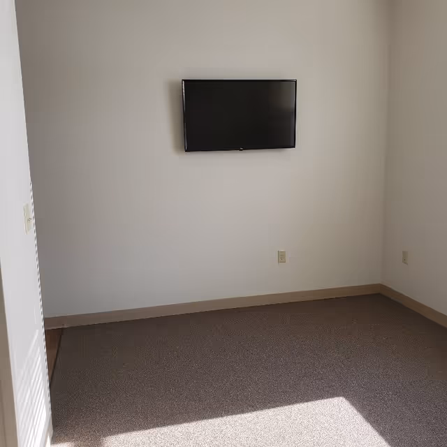 Empty carpeted room with a wall-mounted television on a plain light-colored wall and visible electrical outlets.