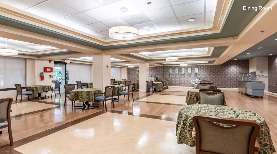 A spacious dining room with multiple tables covered in green patterned tablecloths and surrounded by chairs. The room features a polished wooden floor with a decorative inlay, recessed ceiling lighting, and a patterned wallpaper on one wall. There is a serving station on the right side and an exit door on the left side of the room.