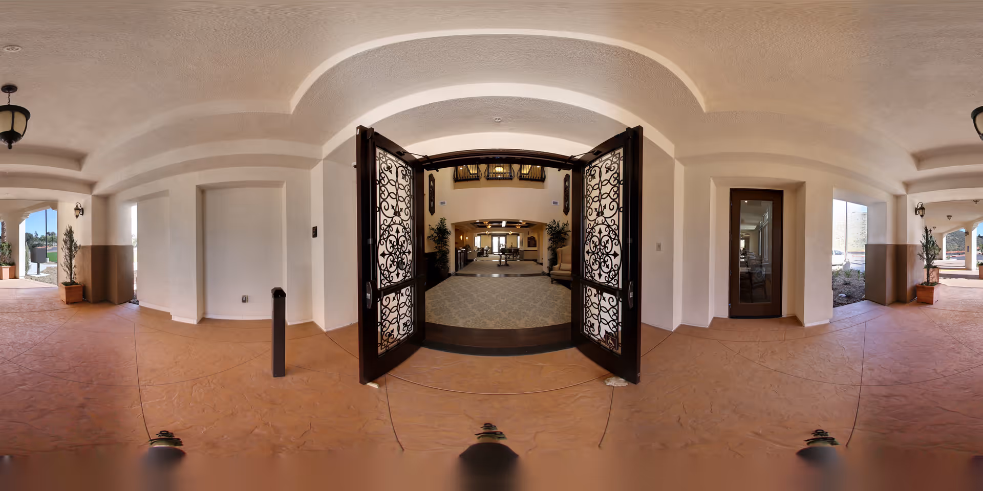 View of the entrance to a senior living facility with ornate double doors open, leading into a spacious interior with carpeted flooring and seating areas. The exterior area has a textured floor and white walls with potted plants and outdoor lighting fixtures.
