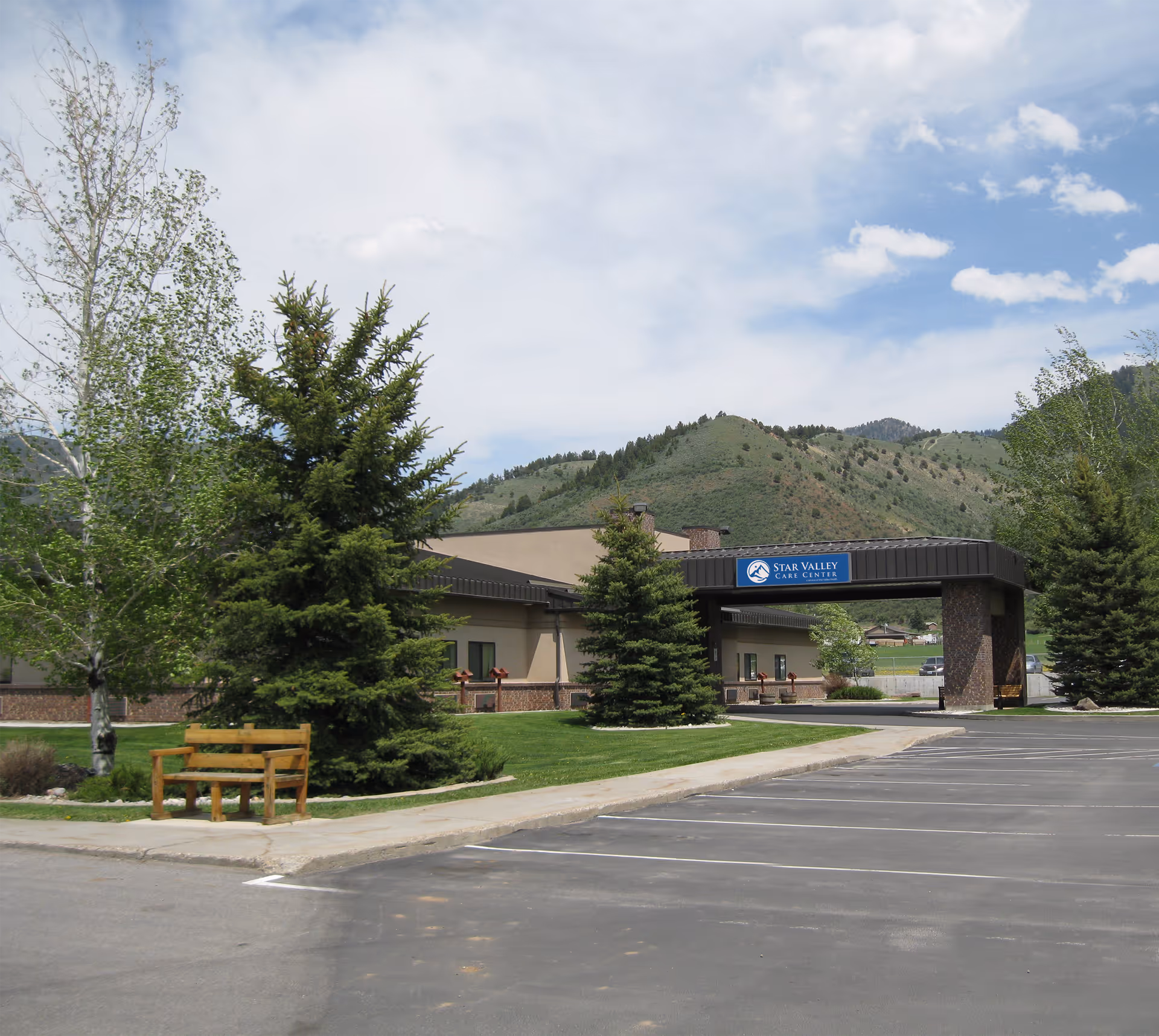 Exterior view of Star Valley Care Center building with a covered entrance, surrounded by green trees and grass, with mountains in the background under a partly cloudy sky. There is a wooden bench near the sidewalk and an empty parking lot in the foreground.