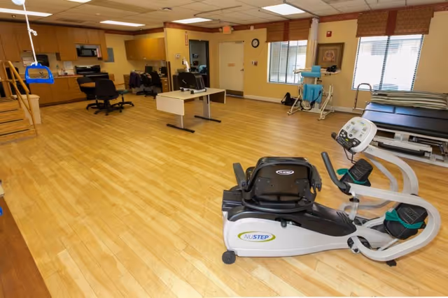 Spacious rehabilitation room with a NuStep recumbent exercise machine in the foreground, therapy tables, chairs, and a kitchenette area in the background.