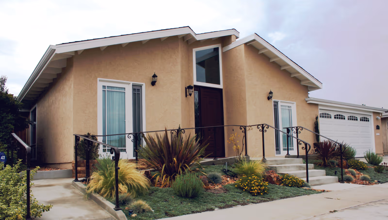 Front exterior of a single-story beige stucco house with a central front door, steps and iron railings, attached garage, and landscaped front yard.