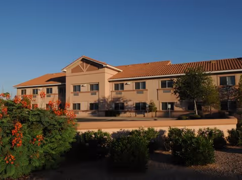 Exterior view of a two-story senior living facility building with a tiled roof, beige walls, and several windows. The foreground features green bushes and a flowering tree with orange blossoms under a clear blue sky.