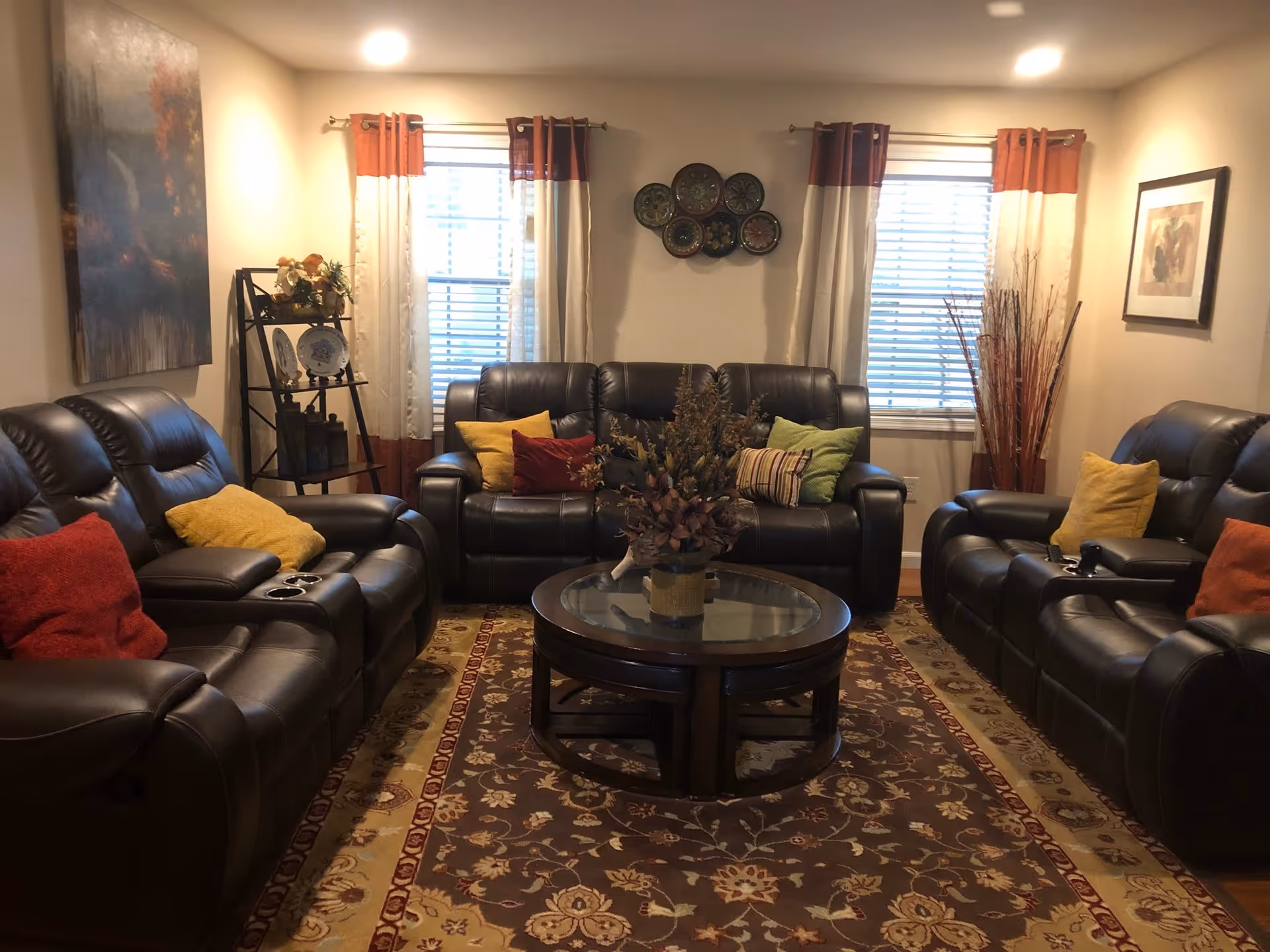 A cozy living room with three dark brown leather sofas arranged around a round glass coffee table with a floral centerpiece. The room has two windows with white and orange curtains, a patterned area rug, and decorative wall art including plates and framed pictures. There is also a small shelving unit with decorative items and a tall vase with dried branches.