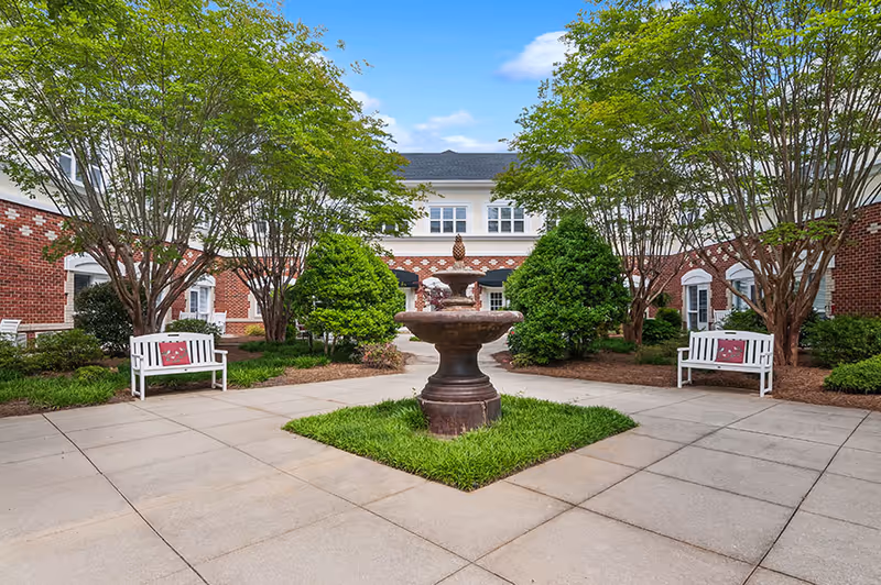 Outdoor courtyard area with a central stone fountain surrounded by green grass and paved walkways. There are two white benches with red cushions on either side, and the courtyard is bordered by trees and bushes. In the background, a two-story brick building with white trim and multiple windows is visible under a blue sky with some clouds.