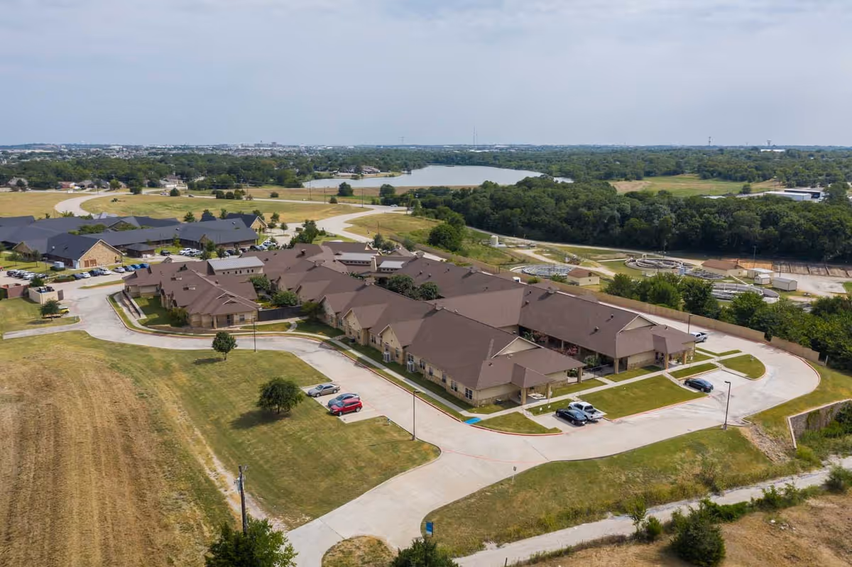 Aerial view of Lakeshore Assisted Living and Memory Care facility showing multiple connected buildings with brown roofs surrounded by green lawns, parking areas with several cars, and a winding driveway. The background includes trees, a small lake, and open fields under a partly cloudy sky.