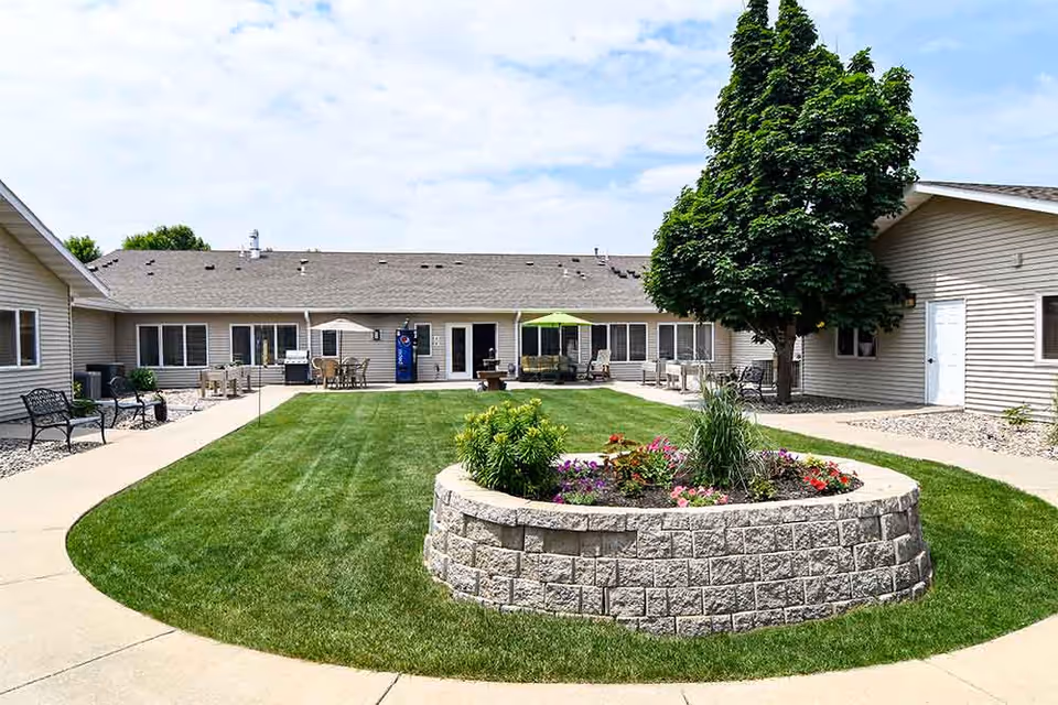 A central courtyard of a memory care facility with a circular raised flower bed, green lawn, benches, patio furniture and single-story buildings around it.