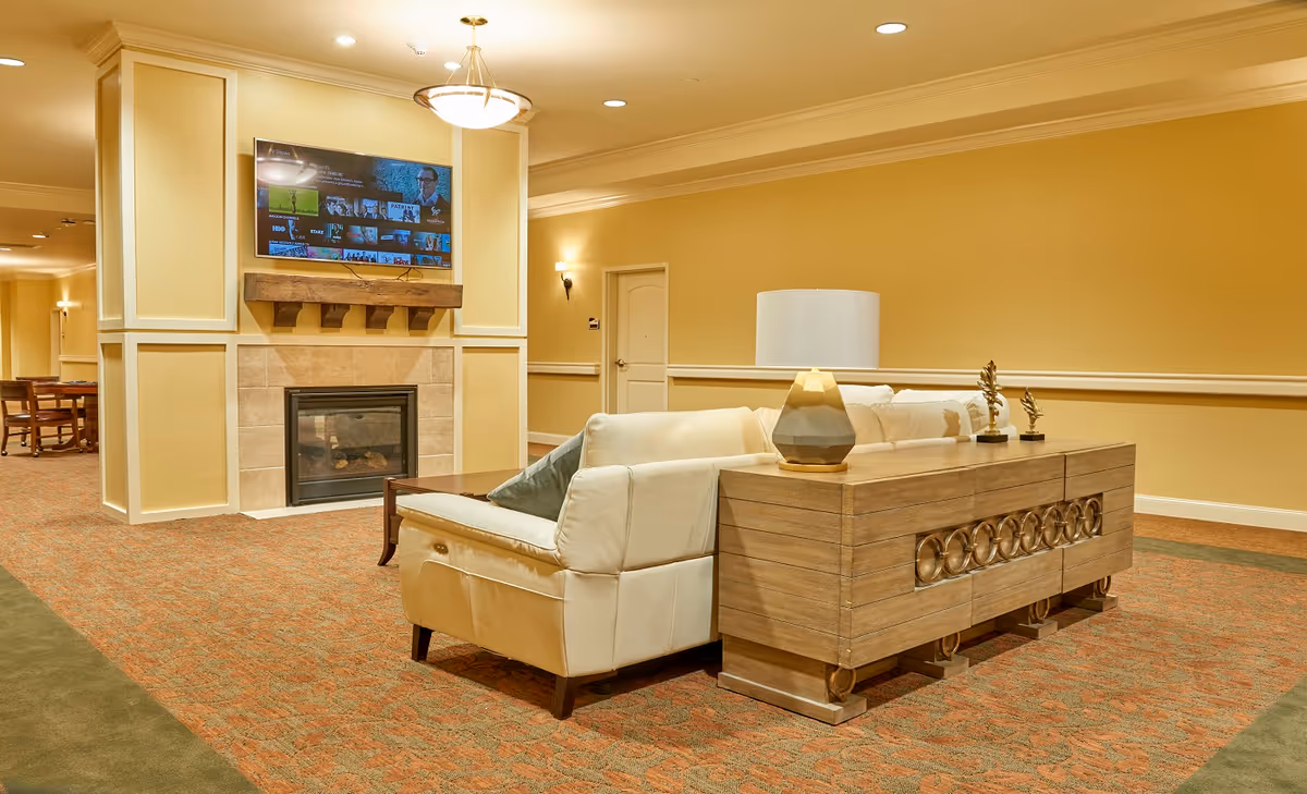 A cozy living room area in a senior living facility featuring a white leather sofa with a gray pillow, a wooden console table with a lamp and decorative items, a fireplace with a wooden mantel, and a wall-mounted flat-screen TV. The room has warm yellow walls, recessed lighting, and carpeted floors with a patterned design.