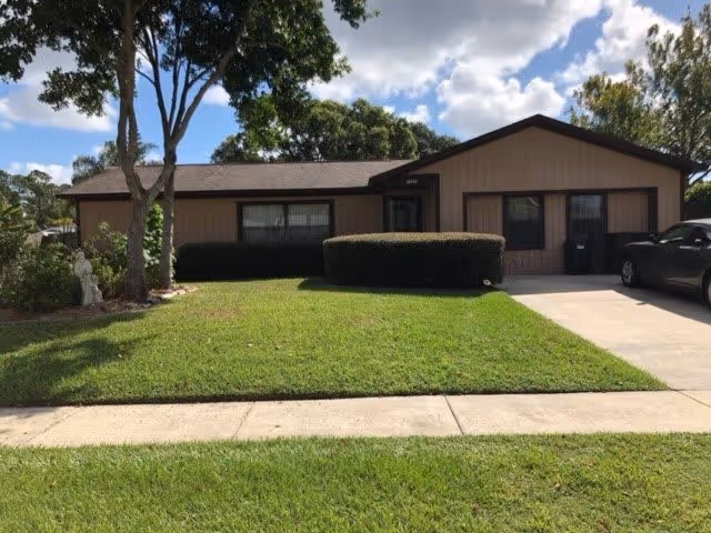 Single-story residential building with beige siding and a dark brown roof, surrounded by a well-maintained lawn and trimmed bushes. A concrete driveway with a parked car is visible on the right side. Trees and a partly cloudy sky are in the background.