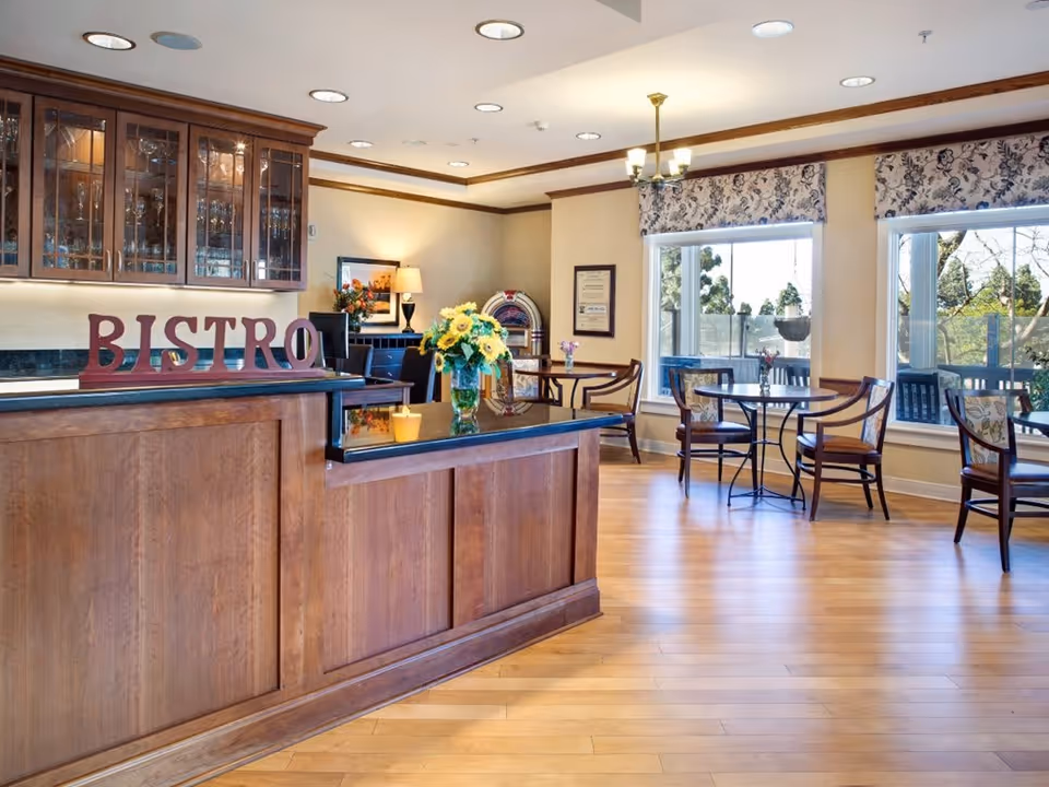 A cozy bistro area inside a senior living facility with wooden floors and furniture. The counter has a sign that reads 'BISTRO' and a vase with yellow flowers. There are glass cabinets with glassware above the counter, and several small round tables with chairs near large windows letting in natural light. The walls are decorated with framed pictures and a lamp, and there is a vintage jukebox in the corner.