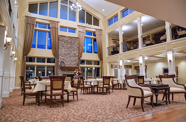 Spacious and elegant dining room with multiple tables and chairs arranged on a patterned carpet. The room features a high ceiling with large windows, tall curtains, a stone fireplace, and a chandelier. There is a second-floor balcony overlooking the area with white columns and additional seating.