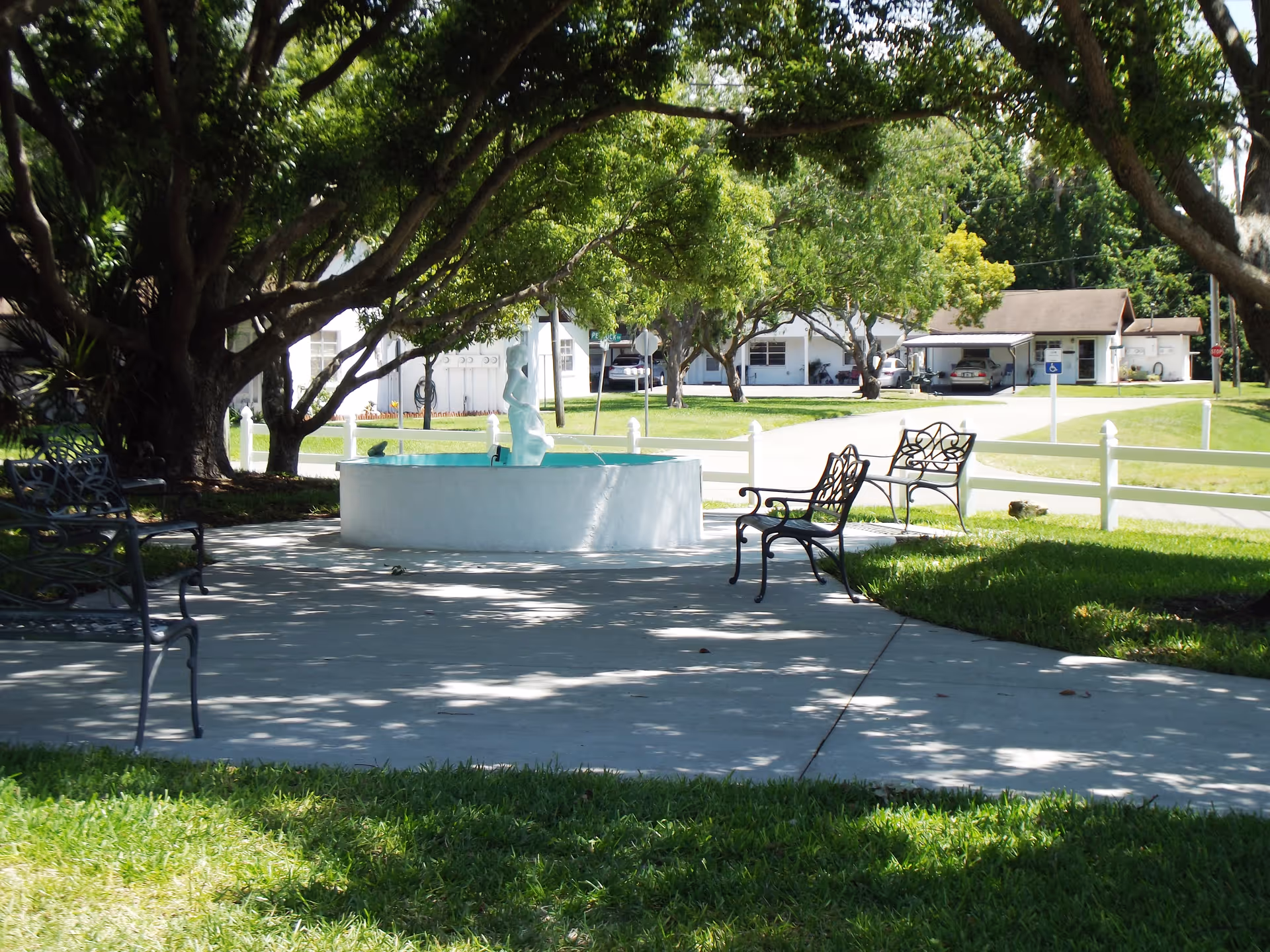 Shaded outdoor courtyard with a central white fountain, metal benches, large trees, and buildings and a white fence in the background.