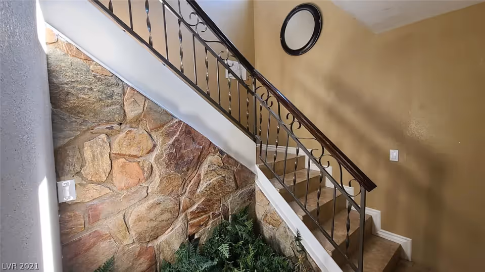 Interior view of a staircase with a decorative wrought iron railing, stone wall on one side, beige painted walls, a round mirror hanging on the wall, and green plants at the base of the stairs.