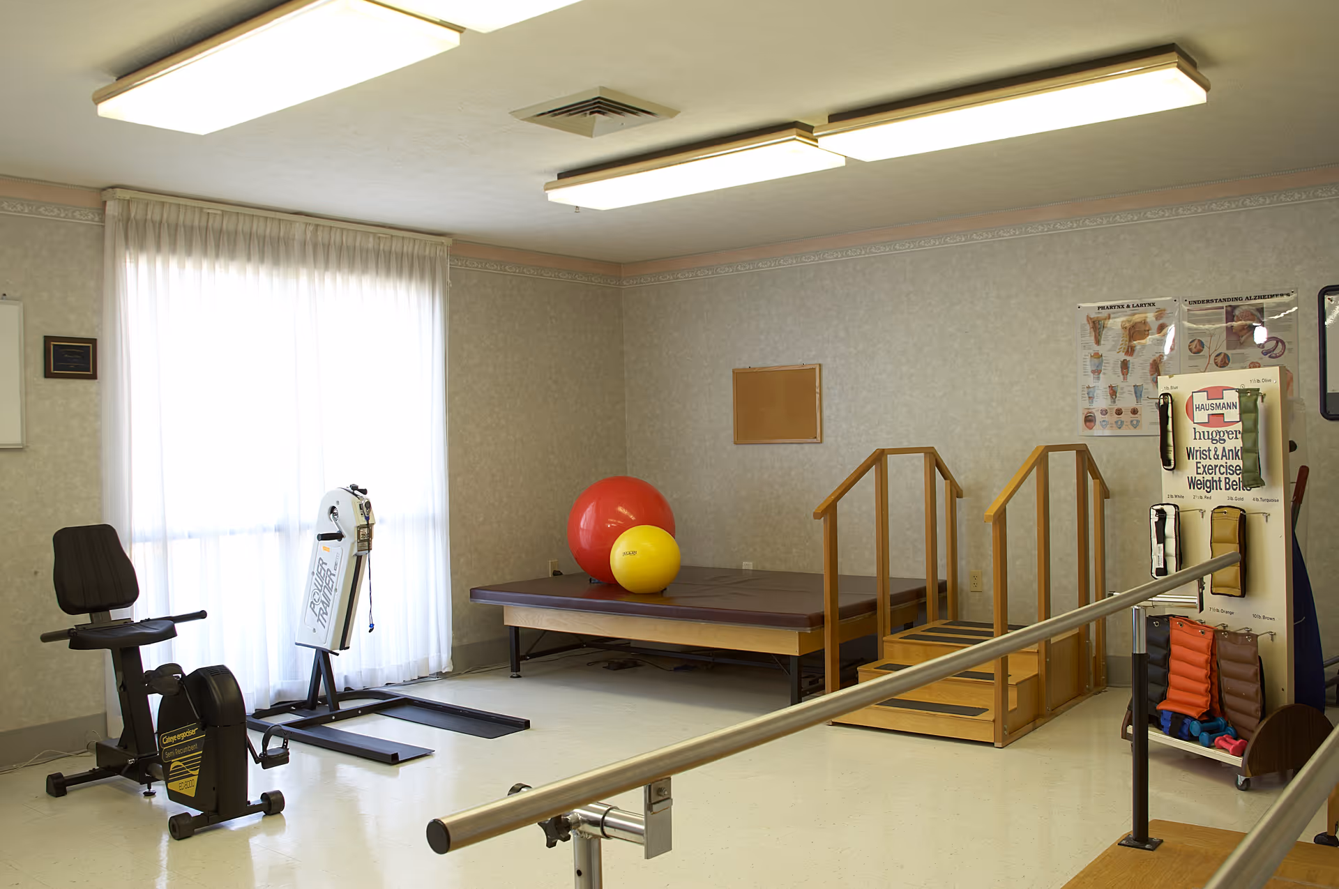 A physical therapy or exercise room with equipment including a stationary exercise bike, a rowing machine, a padded platform with large exercise balls, wooden stairs with handrails, and parallel bars for walking exercises. The room has fluorescent ceiling lights, a window with sheer curtains, and posters on the wall related to exercise and anatomy.