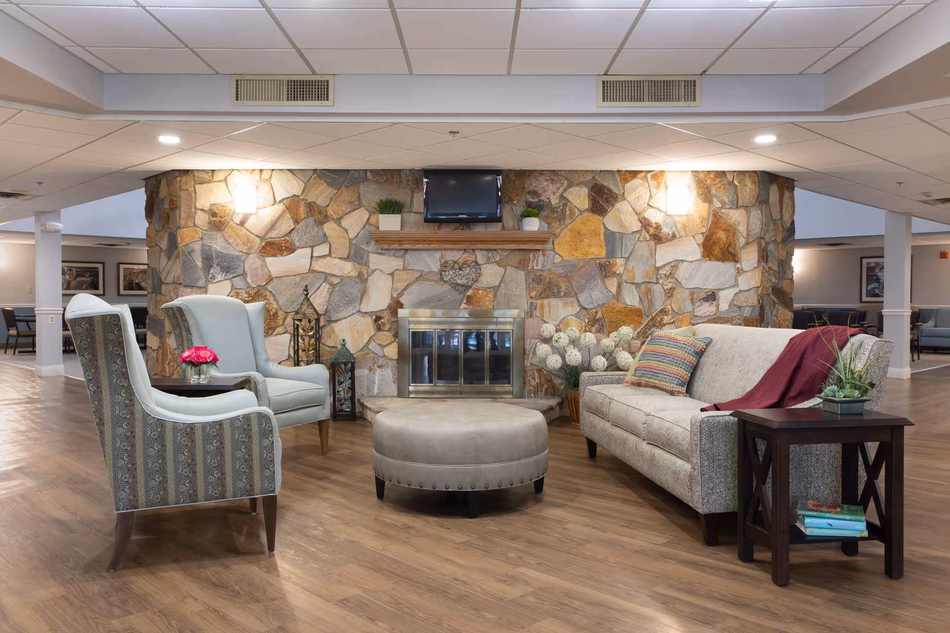 A cozy seating area in a senior living facility featuring a stone fireplace with a mounted TV above it. The area includes a beige sofa with colorful pillows and a red throw, two patterned armchairs, a round ottoman, and a small side table with a plant and books. The room has wooden flooring and soft lighting.