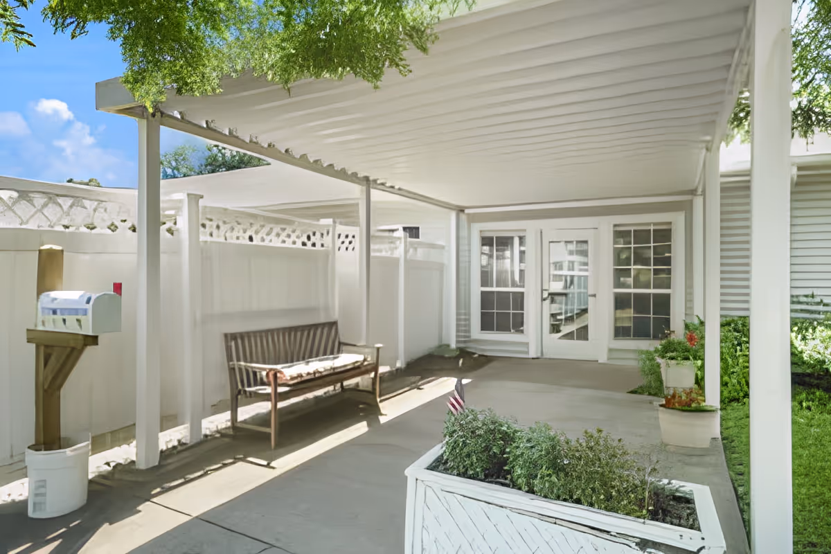 Covered outdoor patio area with a wooden bench, white privacy fence, mailbox on a wooden post, and planter boxes with greenery. The patio has a white pergola roof and leads to a building with glass doors and windows.