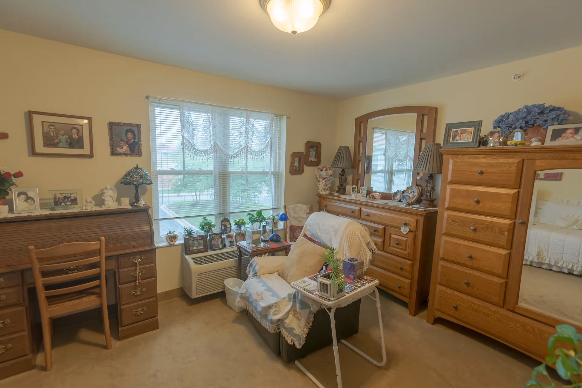 A cozy bedroom with beige walls and carpet, featuring a wooden dresser with a large mirror, a wooden chest of drawers with a mirror, a small armchair covered with a blanket and pillows, a small table with plants and photos, a wooden desk with a chair, and a window with lace curtains letting in natural light. The room is decorated with family photos and various decorative items.
