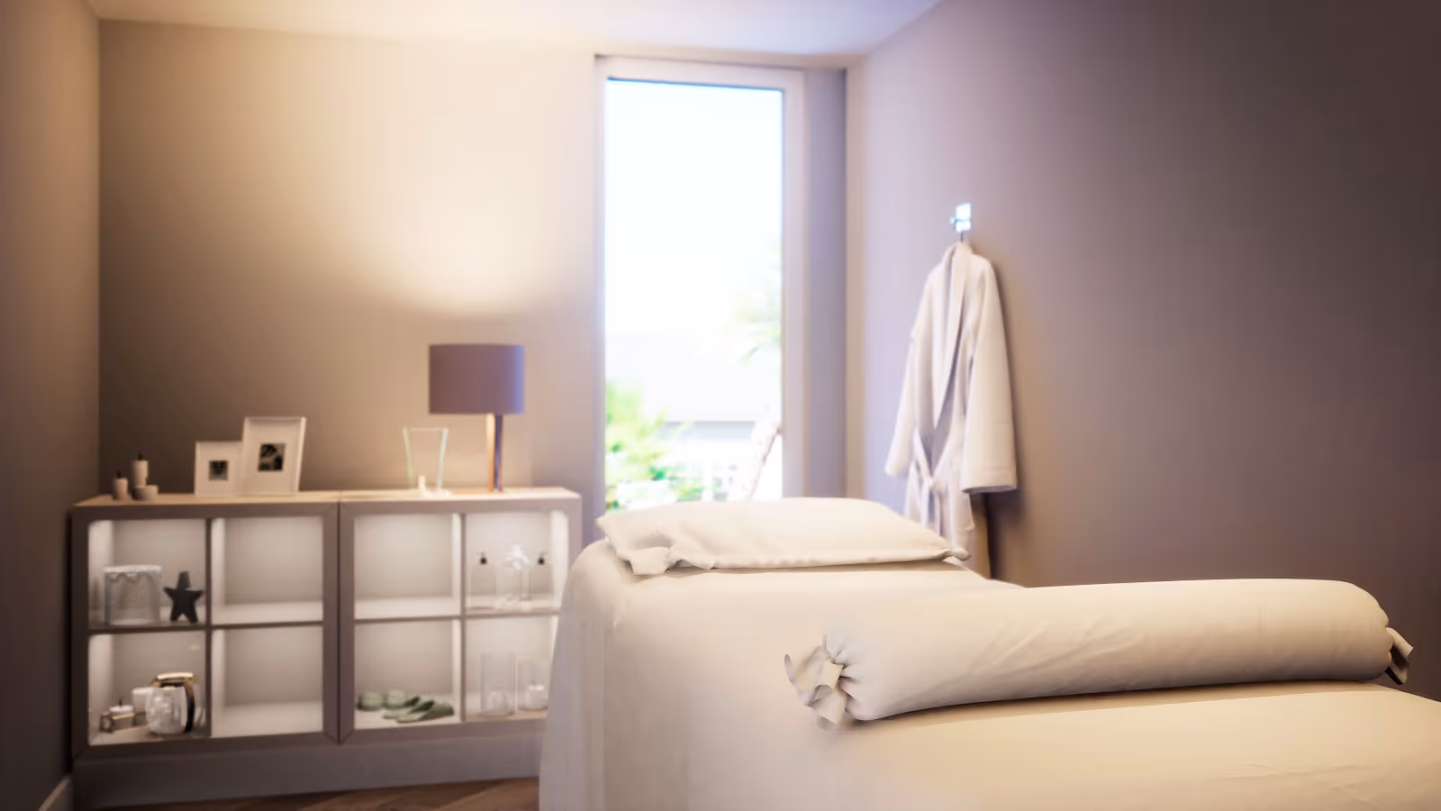 A serene and softly lit room featuring a massage or treatment table covered with white linens and a bolster pillow. Behind the table is a shelving unit with decorative items, a lamp, and framed photos. A white robe hangs on the wall near a tall window letting in natural light.
