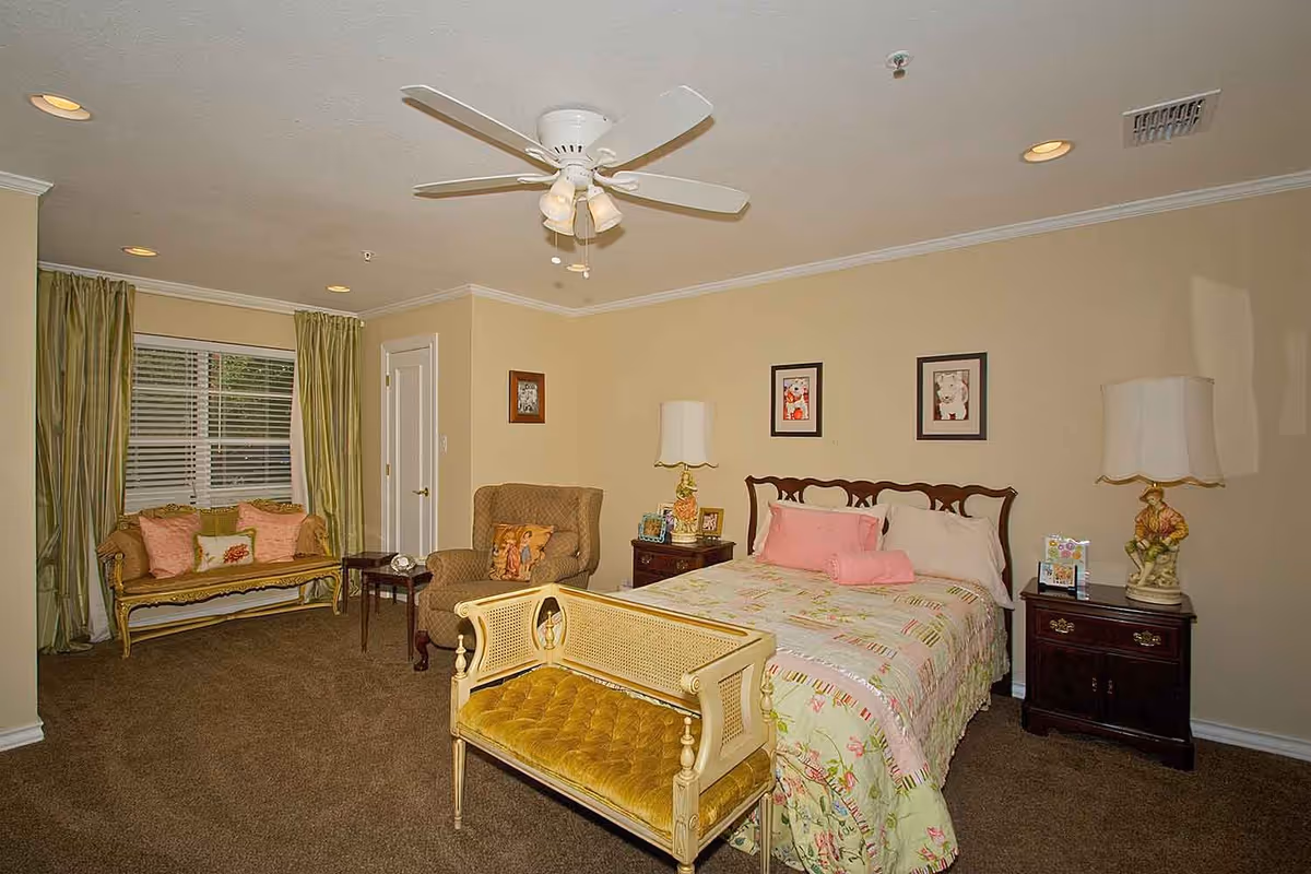 A cozy bedroom with a bed covered in a floral quilt and pink pillows, flanked by two dark wooden nightstands each with a lamp. There is a vintage-style bench at the foot of the bed, a comfortable armchair with a decorative pillow, and a small wooden table. A window with green curtains and a cushioned bench underneath is on the left side of the room. The ceiling has a white fan with lights, and the walls are painted beige.