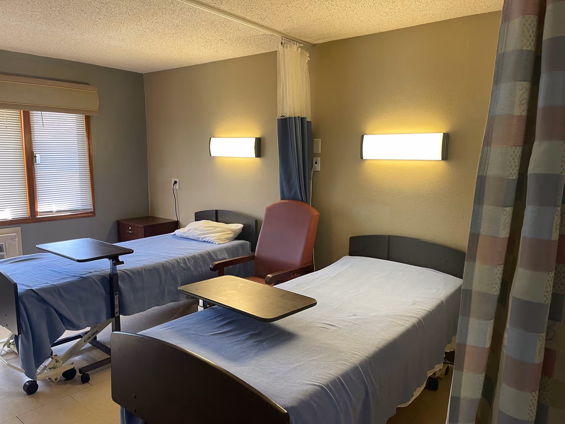 A room with two hospital-style beds covered with light blue sheets, each equipped with an overbed table. There is a red armchair between the beds, wall-mounted lights above each bed, a window with blinds on the left, and a privacy curtain partially visible on the right.